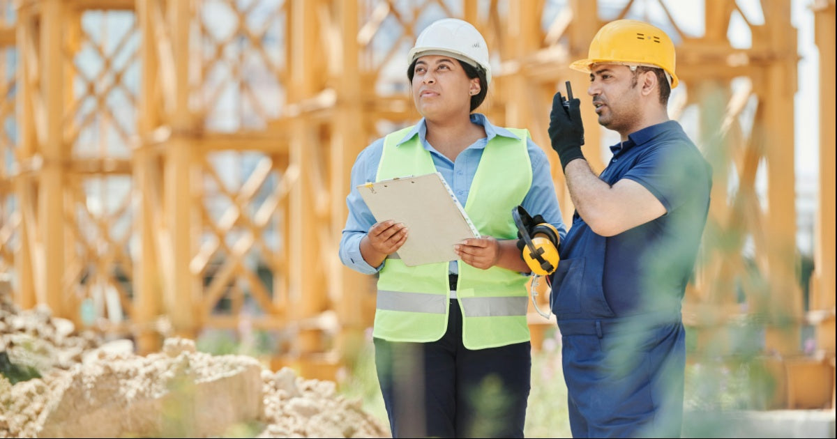 Two construction workers communicating on a job site; one wearing a yellow safety vest and white hard hat holding a tablet, the other wearing a yellow hard hat using a walkie-talkie, with wooden scaffolding in the background.